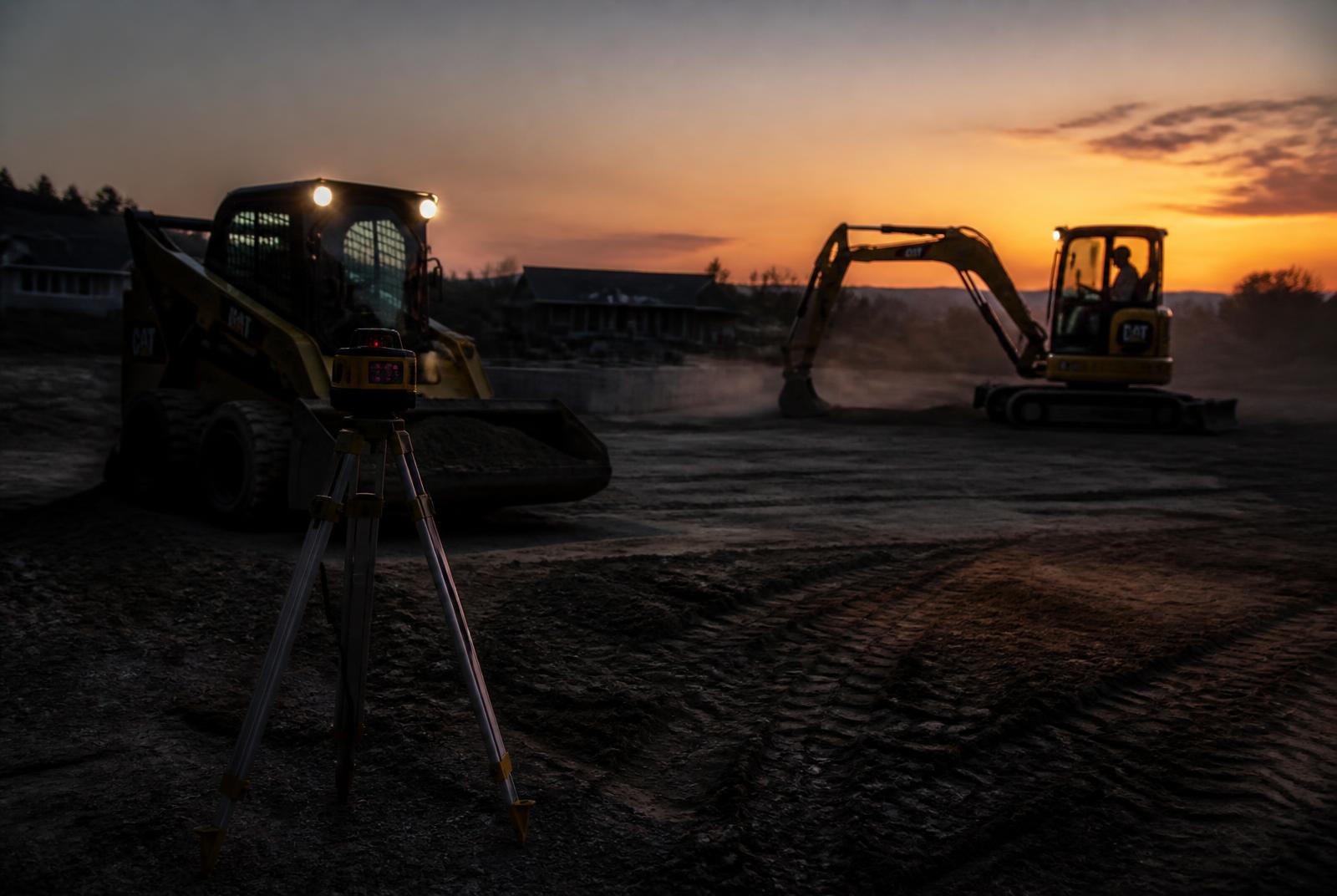 Skid steer and mini excavator grading a pad at dusk with a laser level in the foreground